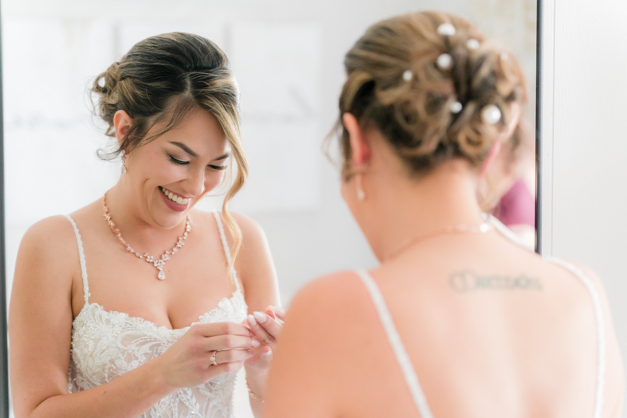 Bride in lace wedding dress smiling at mirror reflection, wearing crystal necklace and updo hairstyle with pearl pins