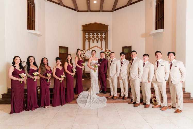 Wedding party photo with bridesmaids in burgundy dresses and groomsmen in cream suits flanking bride and groom under Gothic arch at The Sanctuary