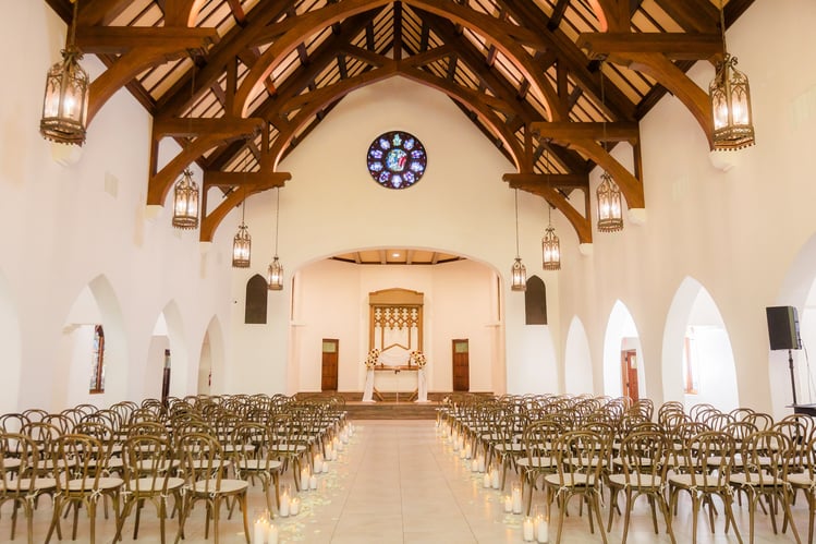 Wedding ceremony setup with wooden cross-back chairs, candlelit aisle, and exposed beam ceiling in Gothic Cathedral