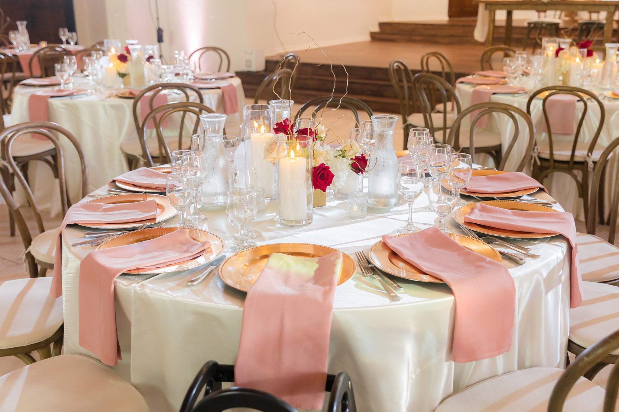 Wedding reception table with white linens, blush pink napkins, gold chargers, and burgundy rose centerpiece at The Sanctuary