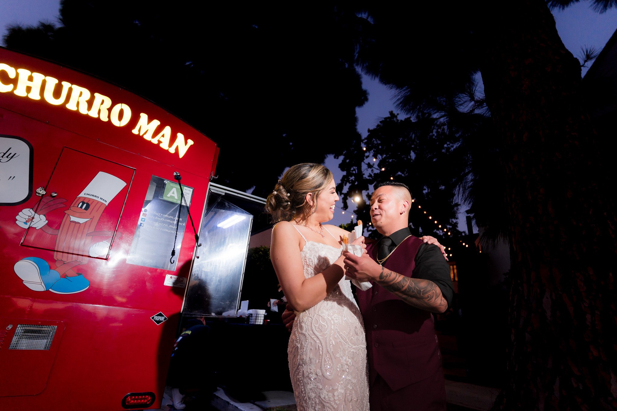 Bride and groom sharing churros in front of red food truck during evening wedding reception