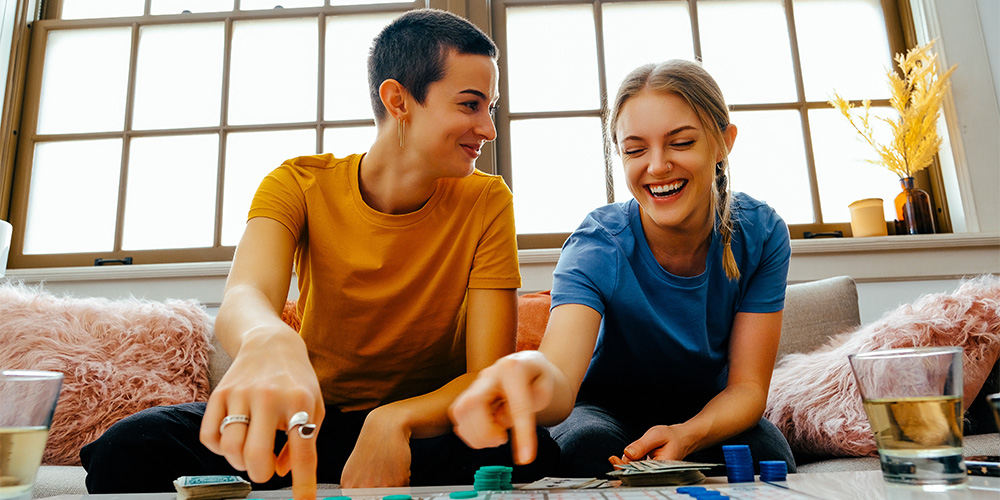 Two young women sitting on couch playing board game with counters, laughing. Happy couple bonding over an indoor leisure activity, smiling and sitting together at a table.