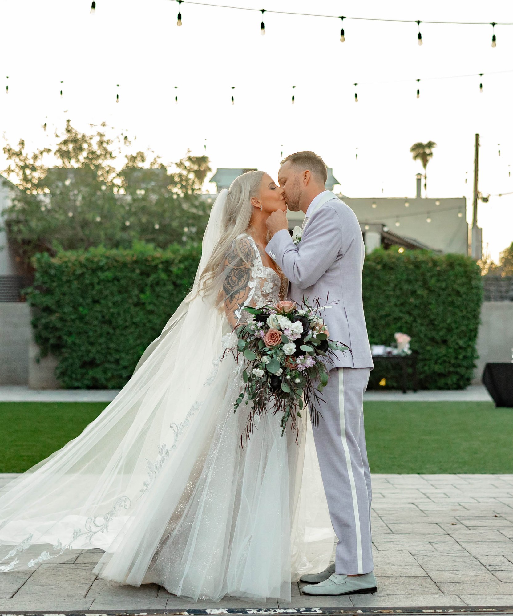 Romantic sunset wedding portrait at Croft Downtown Phoenix, bride in lace dress and groom in light gray suit with floral arrangements and string lights