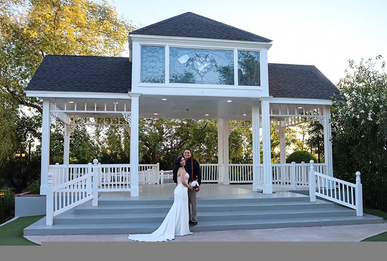 Bride and groom standing on the steps of a charming white gazebo at Lindsay Grove, surrounded by lush greenery. The bride wears a fitted white gown with a long train, holding a bouquet of red and white flowers, while the groom smiles beside her in a black shirt and beige pants