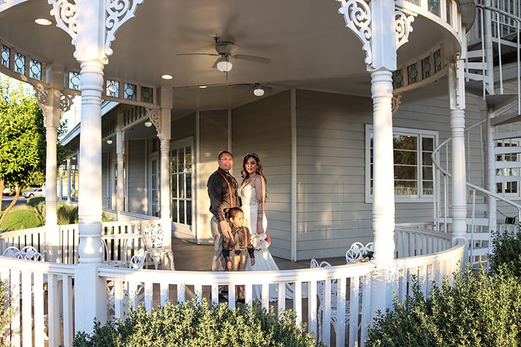 The joyful wedding couple stands on the porch of a Lindsay Grove, accompanied by their young son. The bride is wearing a fitted white gown, while the groom is dressed in a black shirt and beige pants. The setting features decorative white railings and lush greenery, creating a beautiful backdrop.