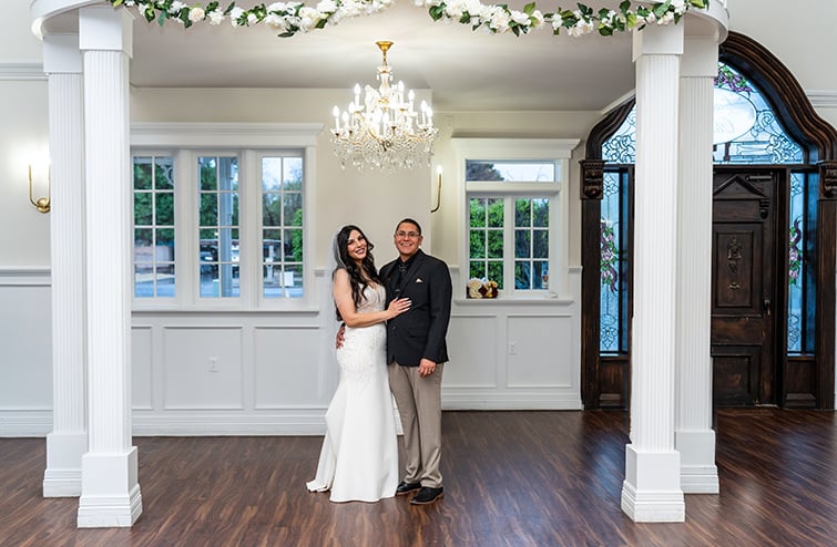 The smiling couple stands together in the beautifully decorated indoor venue, with the bride wearing a white fitted wedding dress and the groom in a dark suit jacket with beige pants. The backdrop features elegant columns, a chandelier, and stained-glass windows.