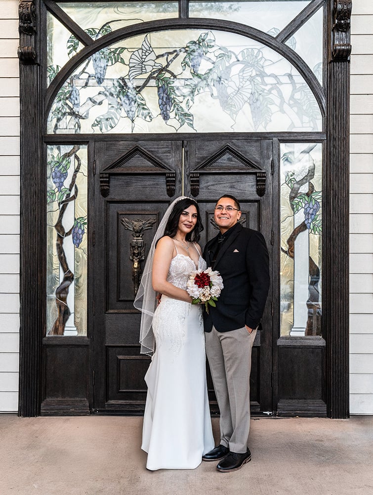 Bride and groom posing in front of an ornate wooden door with stained glass accents at Lindsay Grove. The bride, in a fitted white gown and veil, holds a bouquet of red and white flowers, while the groom wears a black blazer and beige pants, smiling warmly beside her.