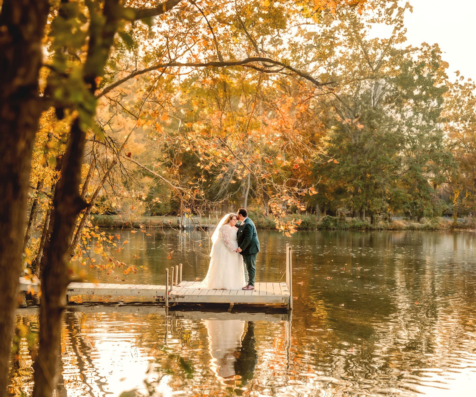 Bride and groom standing on wooden dock surrounded by stunning fall foliage with golden light reflecting on the water at Riverwood Manor - Golden Clover Photography