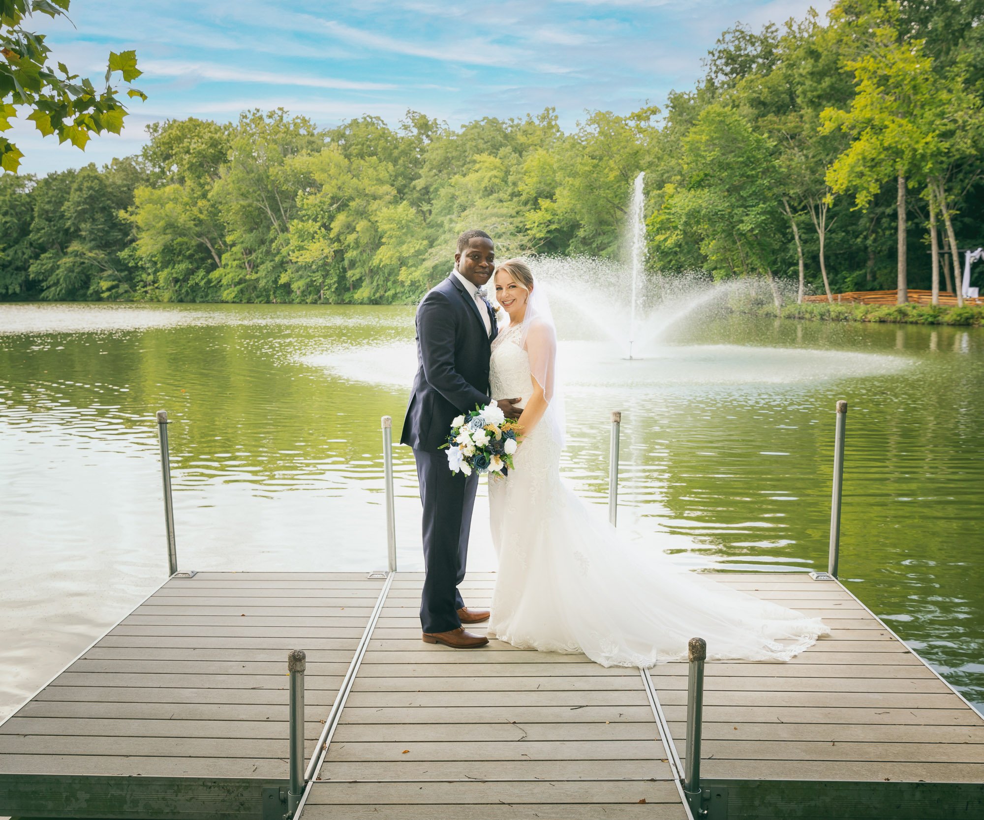 Newlywed couple posing on wooden dock at Riverwood Manor with fountain and lush green trees creating a romantic lakeside backdrop - Ken Thomas Photography