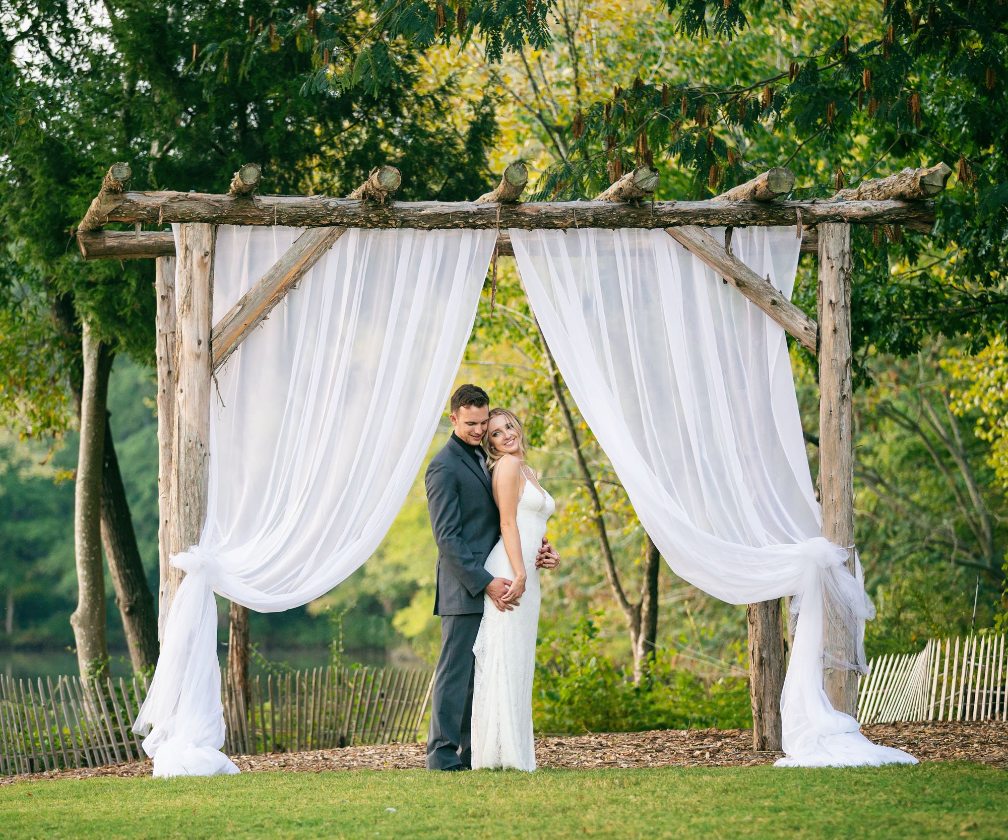 Newlyweds posing by rustic wooden pergola with white sheer drapery at Riverwood Manor's woodland ceremony site, surrounded by lush greenery and overlooking the lake in Harrisburg, NC - Ken Thomas Photography