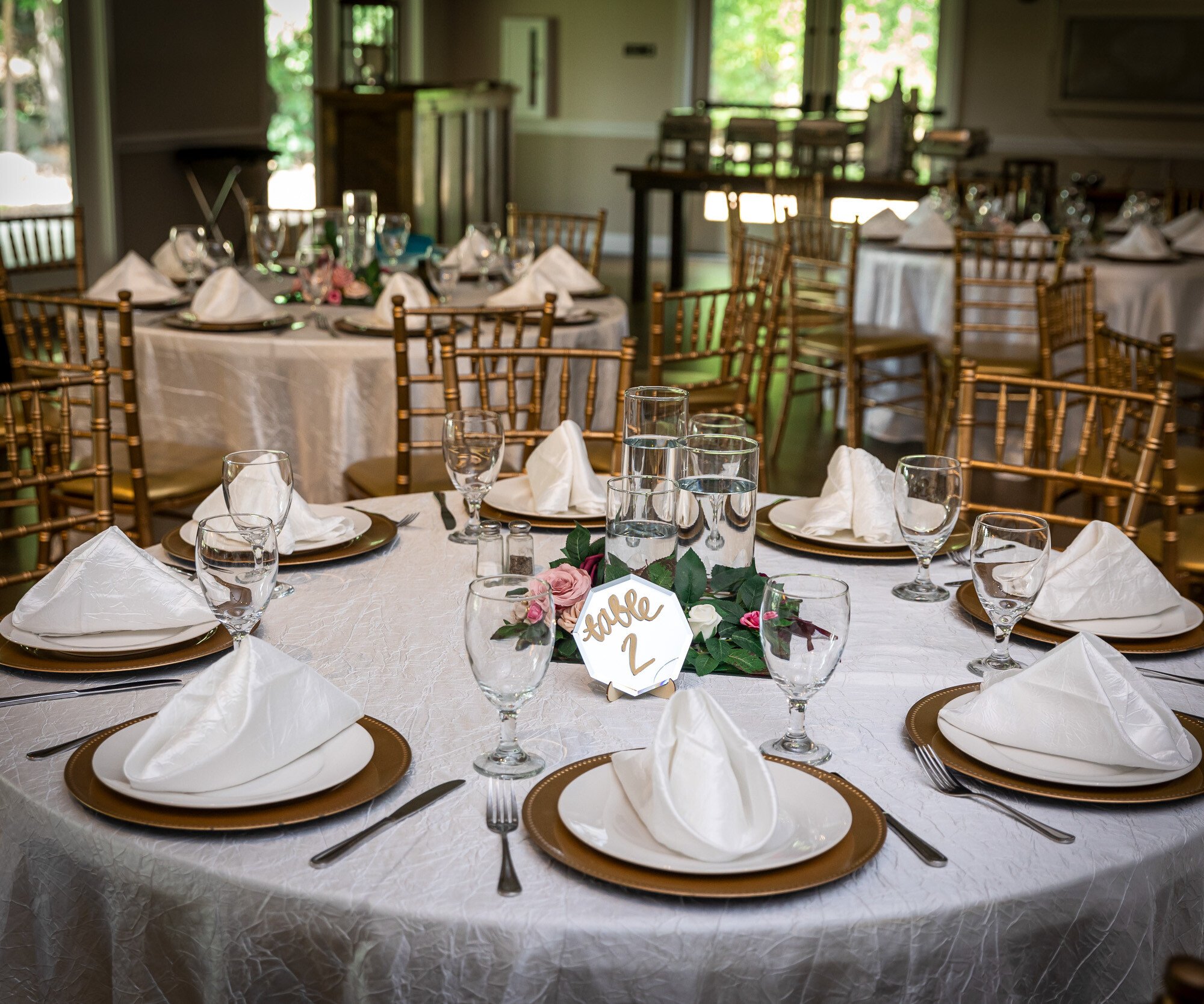 Detailed view of elegant wedding reception table at Riverwood Manor featuring white linens, gold charger plates, folded napkins, crystal glassware, gold chiavari chairs, and a table number with floral accent in the intimate dining space in Harrisburg, NC - Ken Thomas Photography