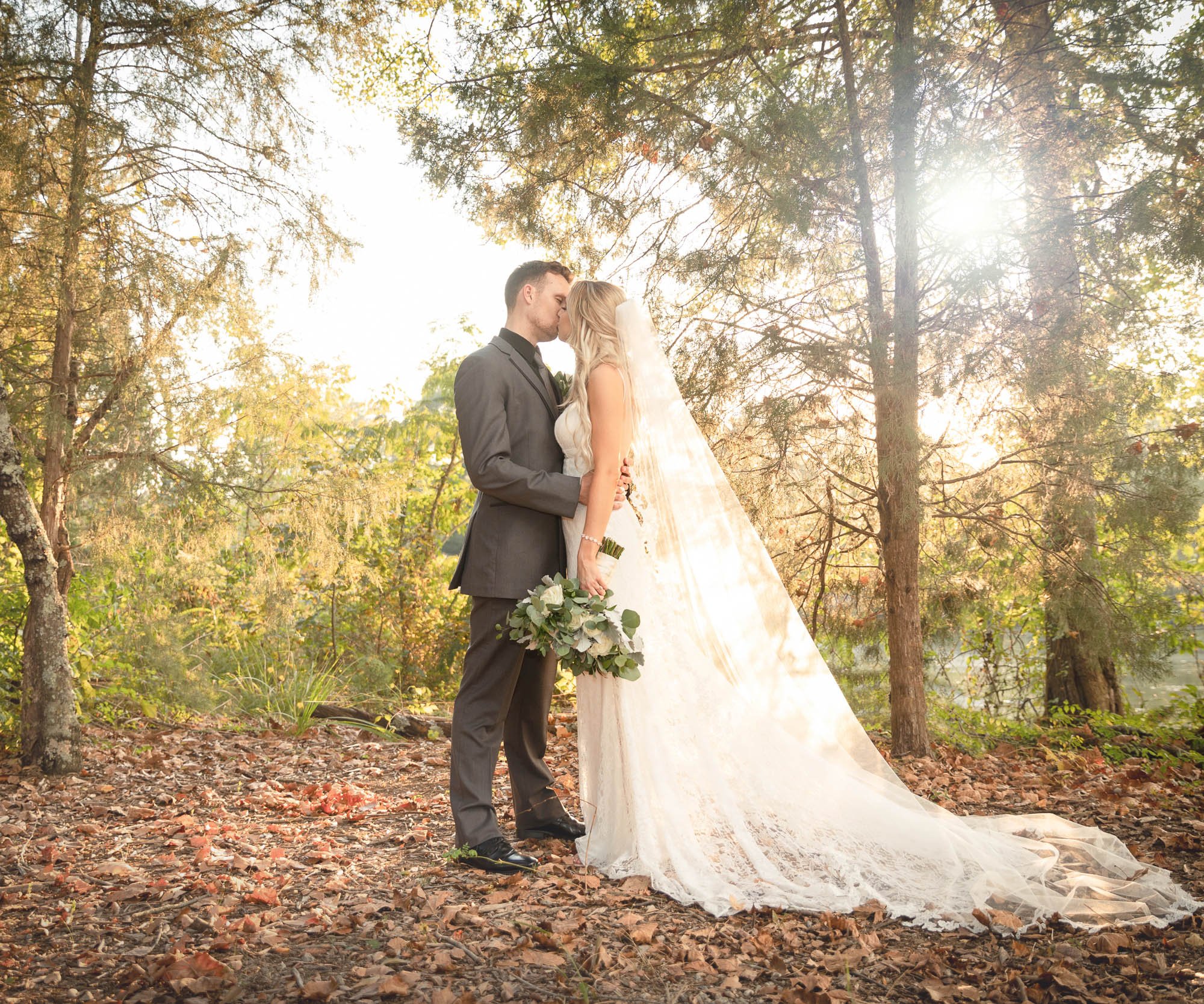 Bride and groom kissing with sunlight filtering through autumn trees at Riverwood Manor, showcasing bride's flowing veil and cathedral train against fall foliage and woodland background - Ken Thomas Photography