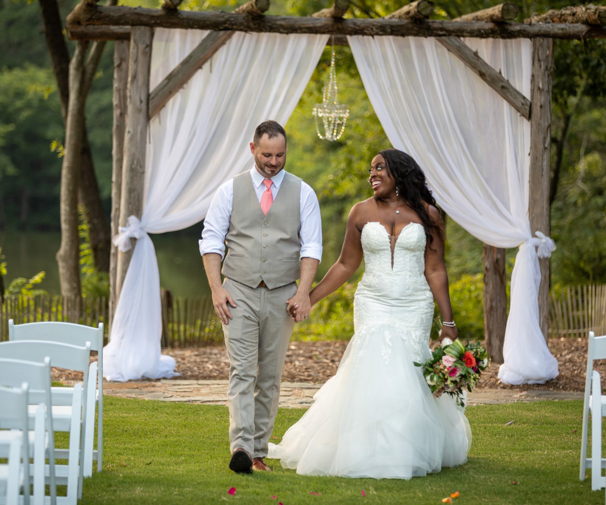 Happy bride and groom walking down the aisle after ceremony at Riverwood Manor, featuring rustic log pergola with white drapery, crystal chandelier, and lakeside background in Harrisburg, NC - Ken Thomas Photography