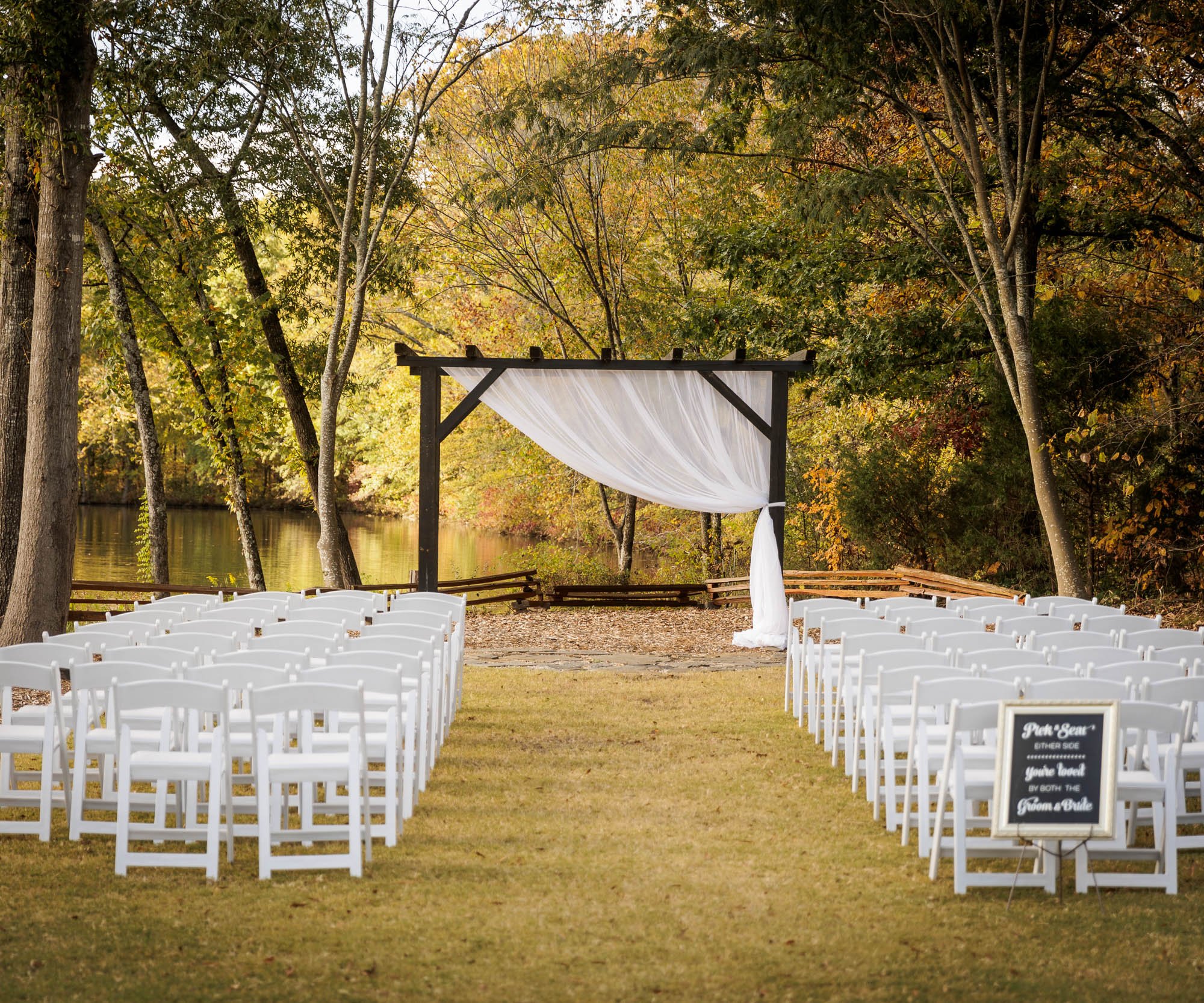 Outdoor wedding ceremony setup at Riverwood Manor with white chairs facing a wooden pergola draped in white fabric overlooking a serene lake with fall foliage - Ken Thomas Photography