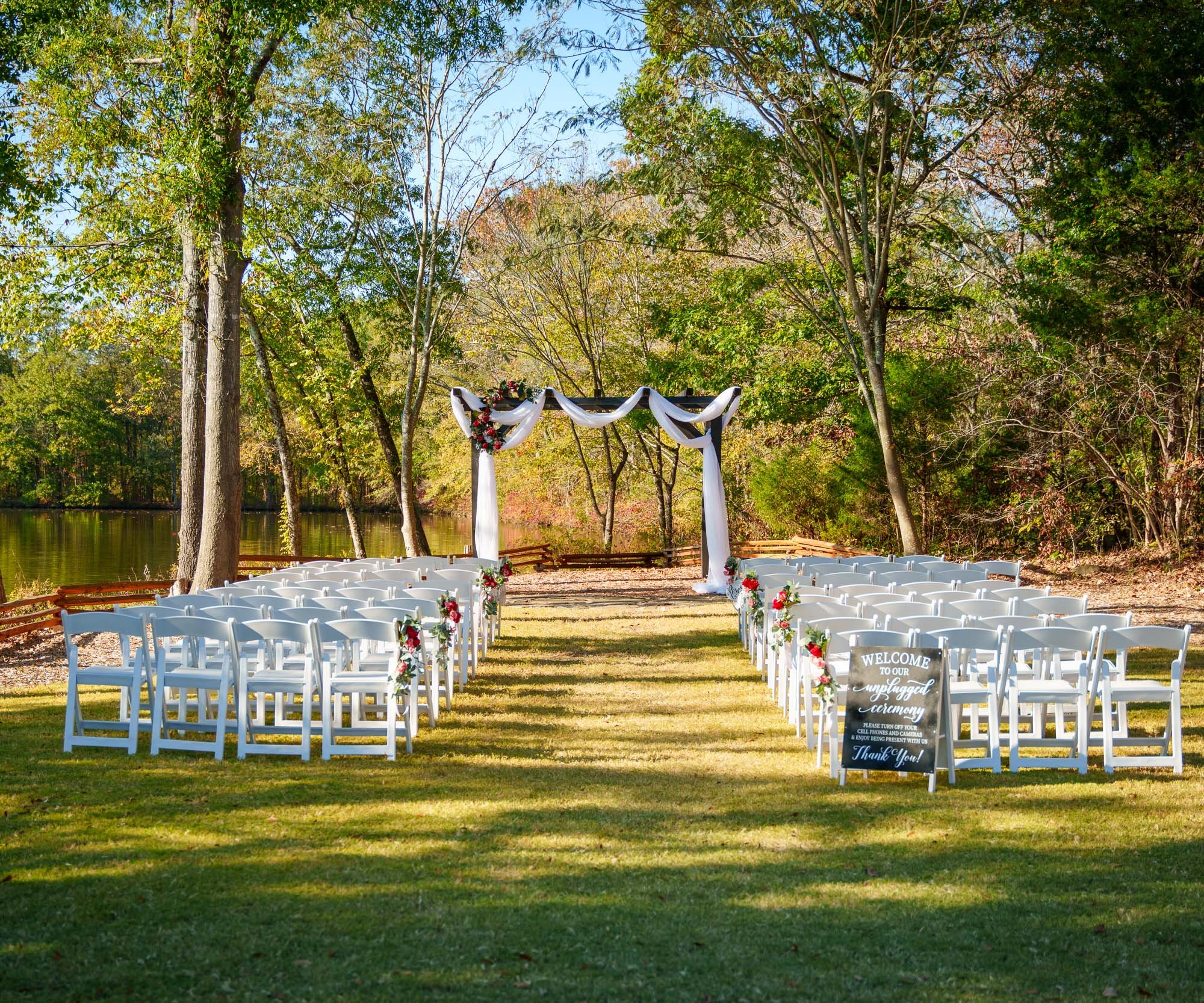 Autumn wedding ceremony setup at Riverwood Manor featuring white chairs, rustic wooden arbor with white drapery, and colorful fall foliage surrounding the lakeside setting - Foxbeau LLC