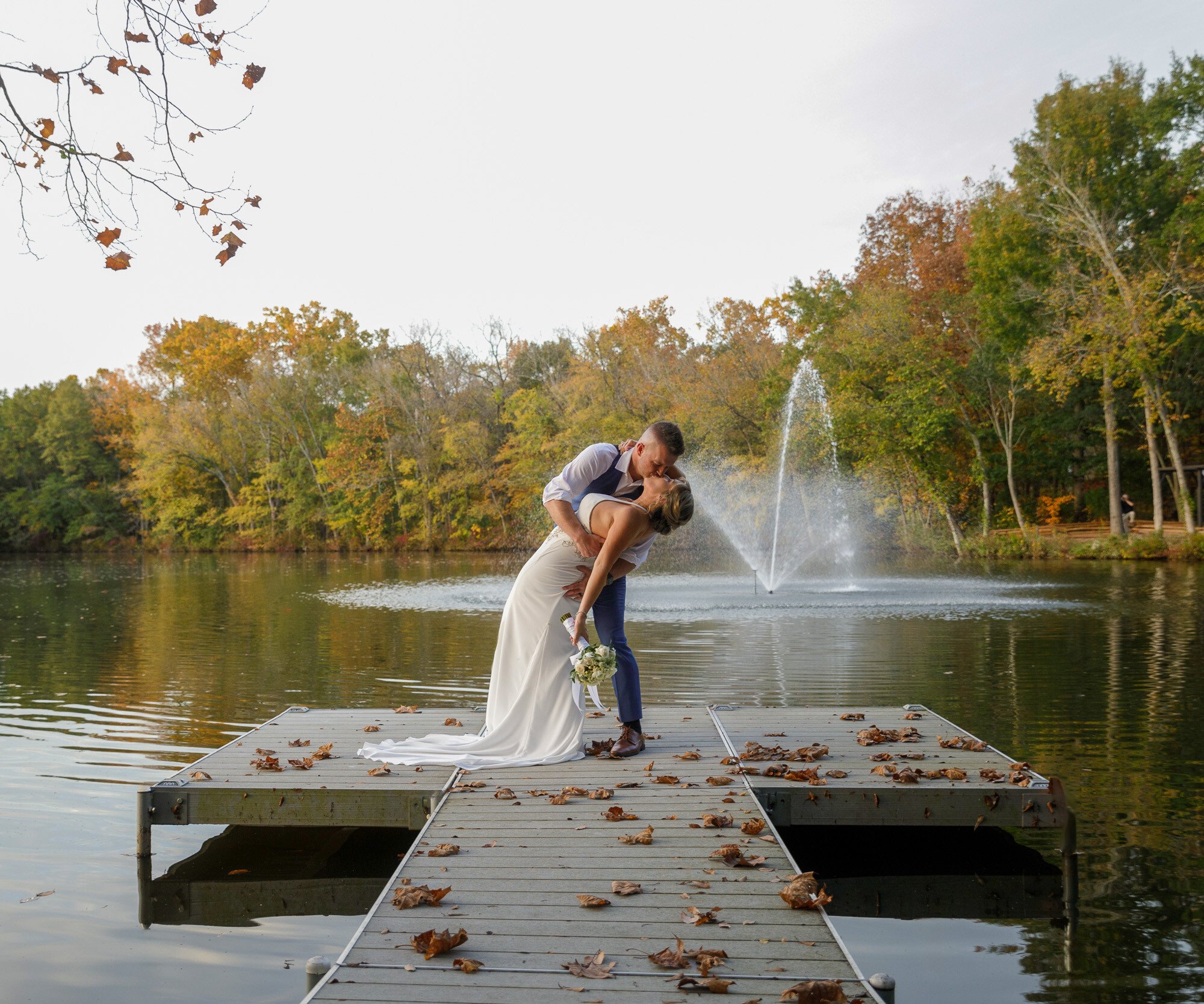 Bride and groom sharing a dip kiss on Riverwood Manor's wooden dock with fall leaves and tranquil lake waters creating a stunning wedding portrait - Ken Thomas Photography