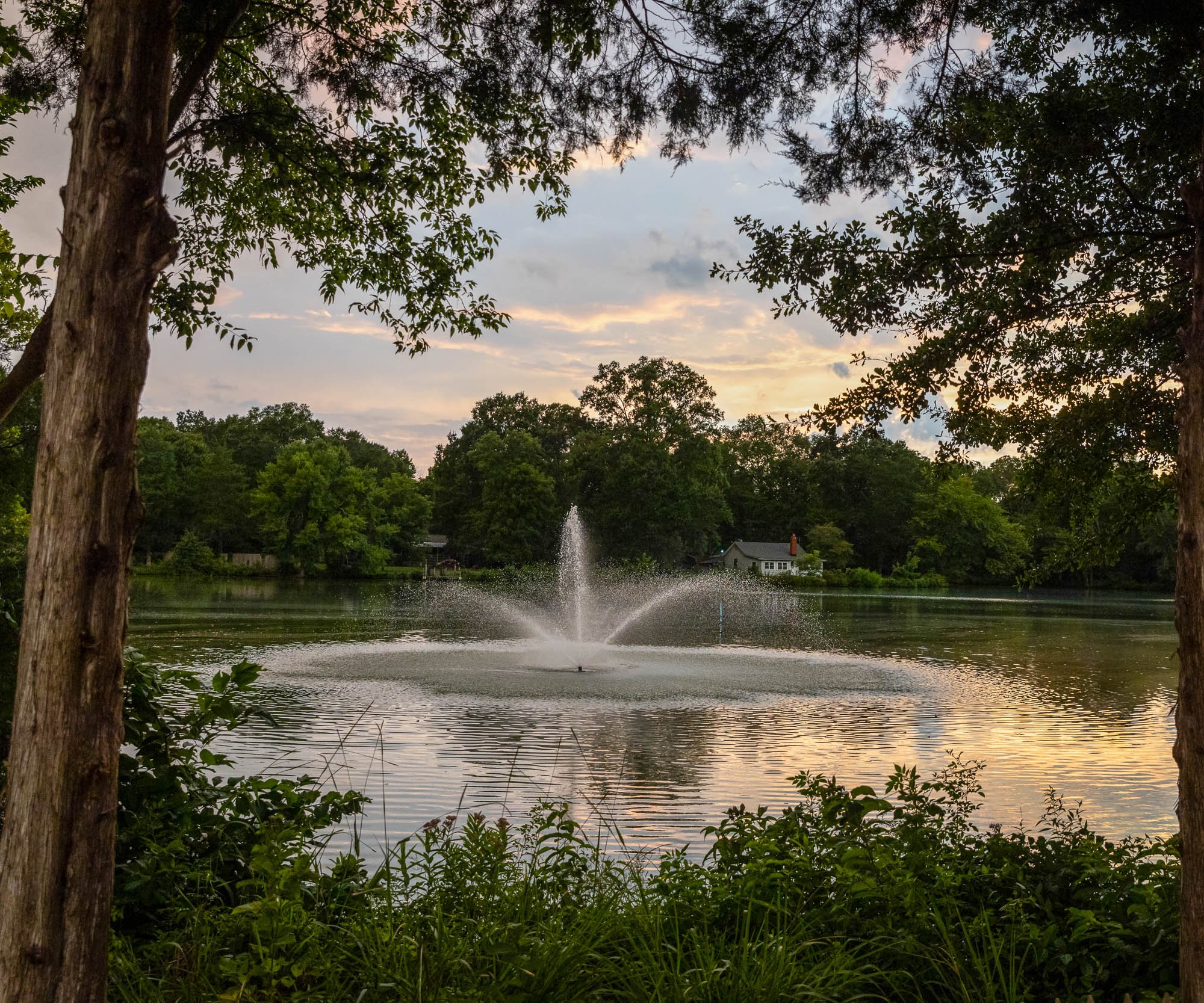 Scenic view of Riverwood Manor's lake with fountain at sunset, framed by mature trees with cottage visible in background, showcasing the venue's serene natural setting in Harrisburg - Ken Thomas Photography