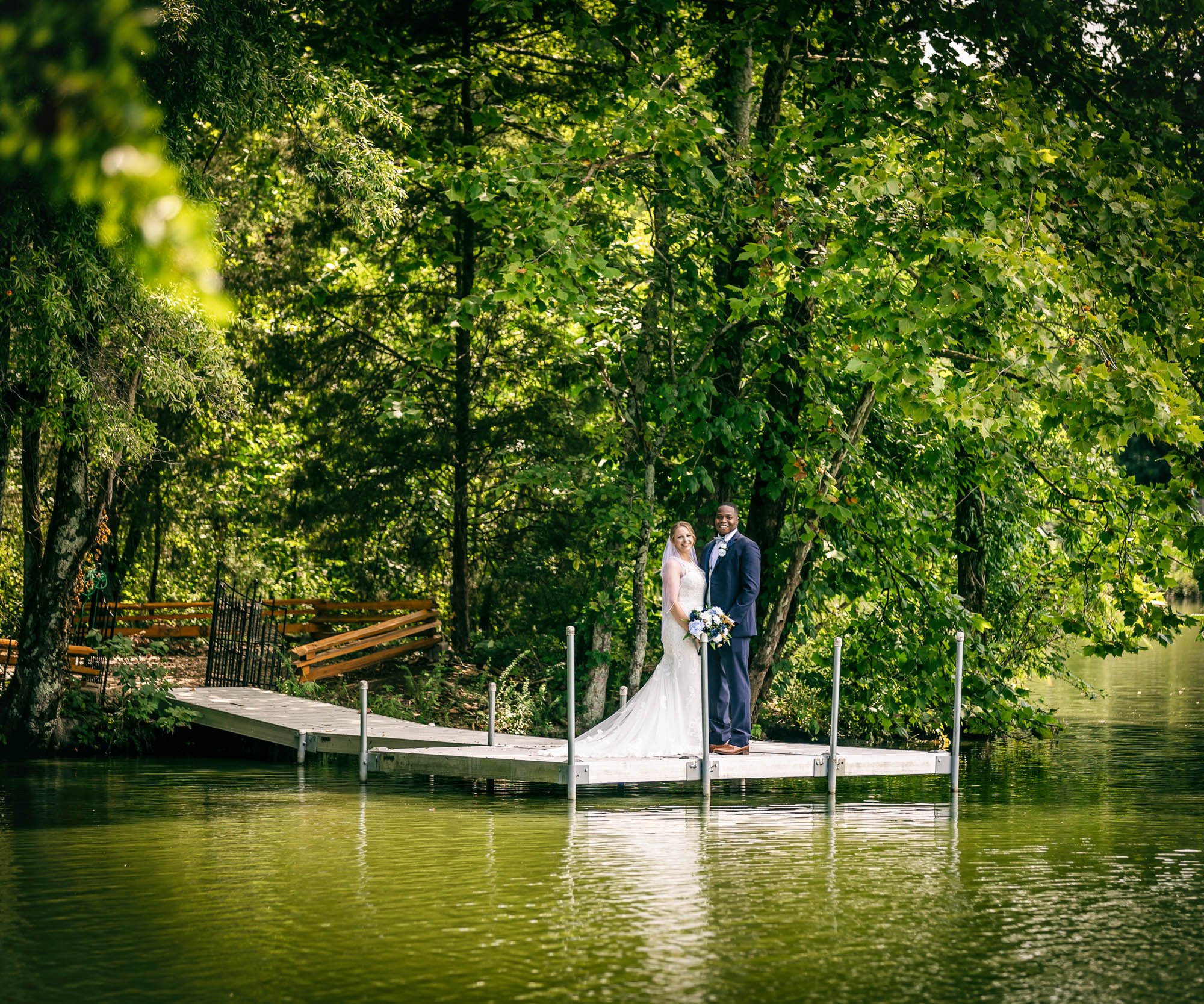 Wedding couple standing on Riverwood Manor's dock surrounded by water with mature trees and woodland setting creating a natural portrait location - Ken Thomas Photography