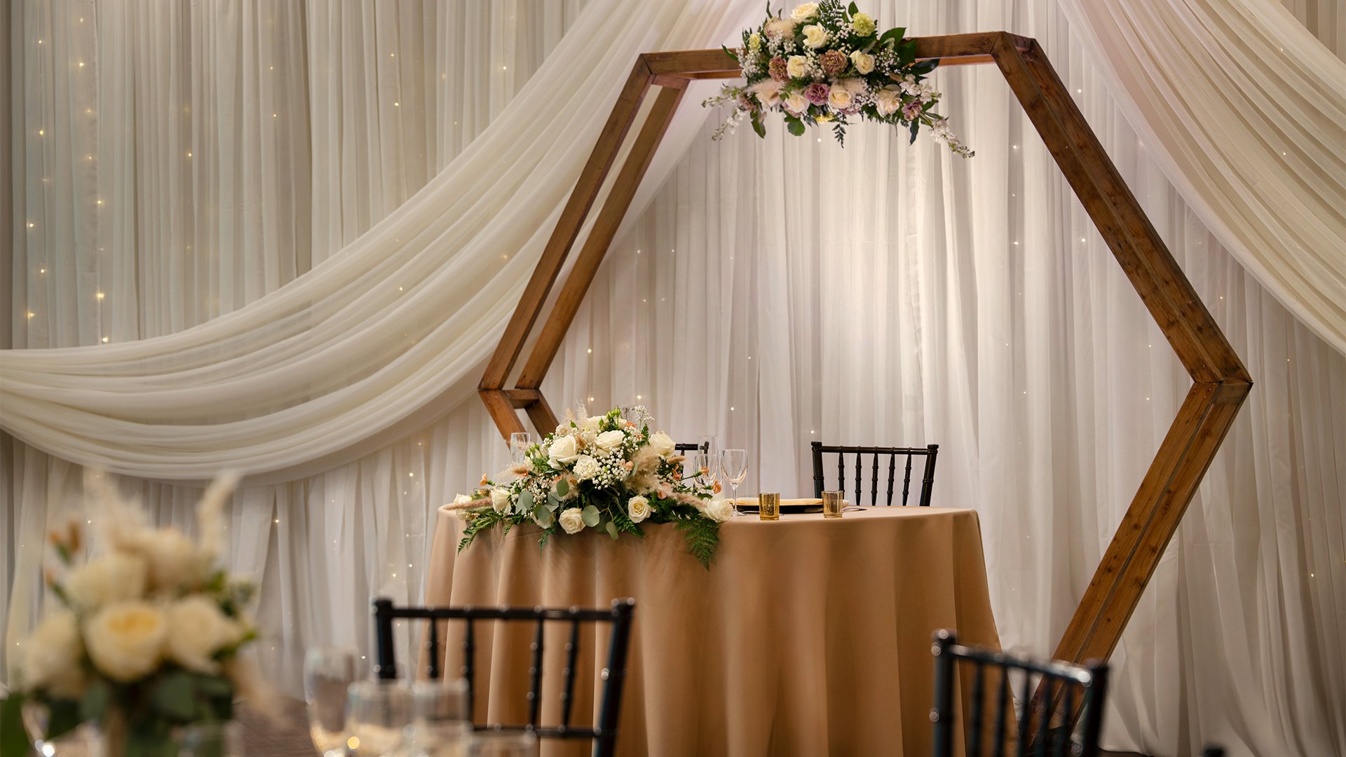 Elegant Wedding Sweetheart Table with Hexagonal Wood Arch, White Draping and Rose Floral Arrangements