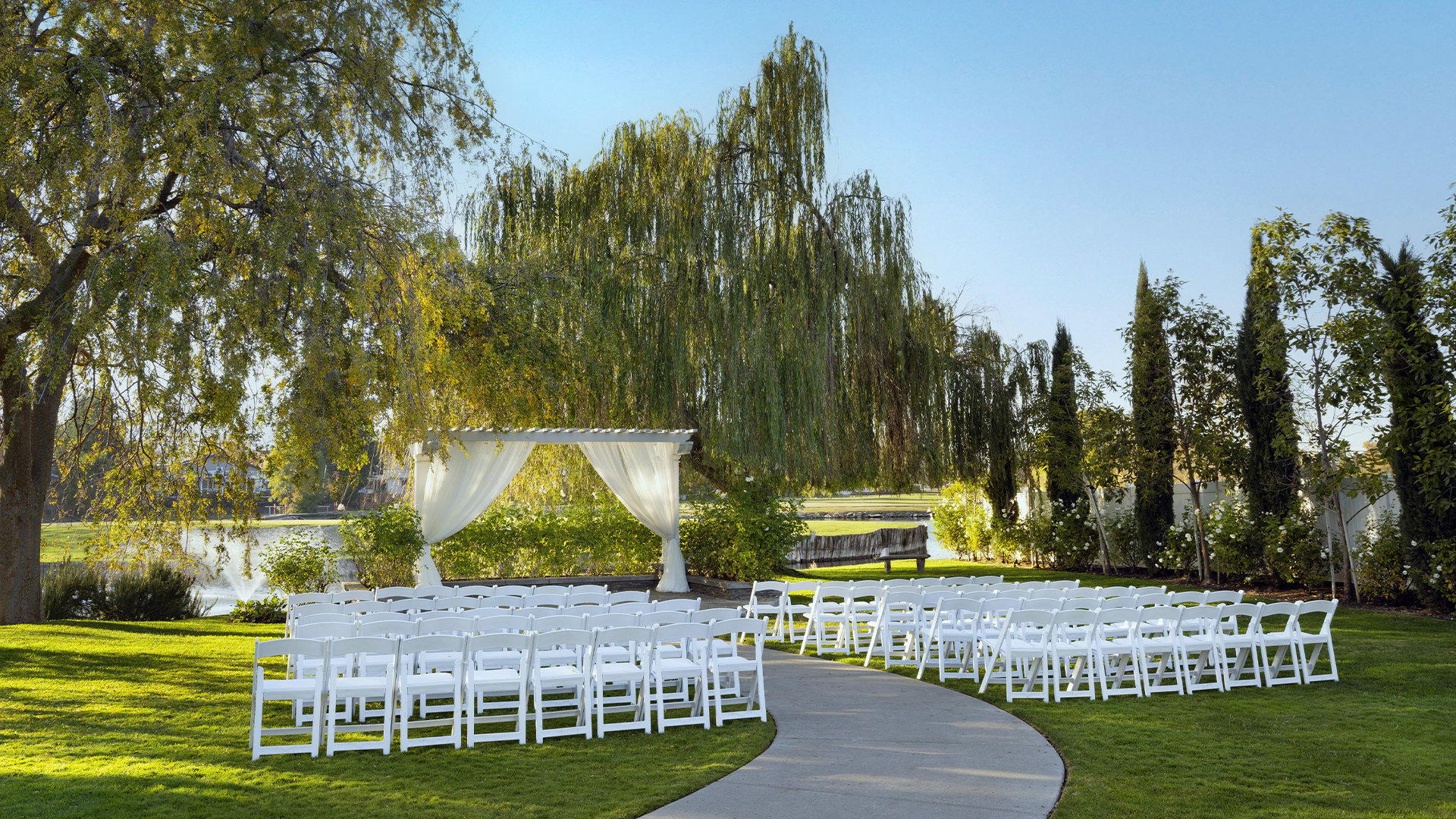 Lakefront Wedding Ceremony Setup with White Garden Chairs and Draped Arch at Sunset