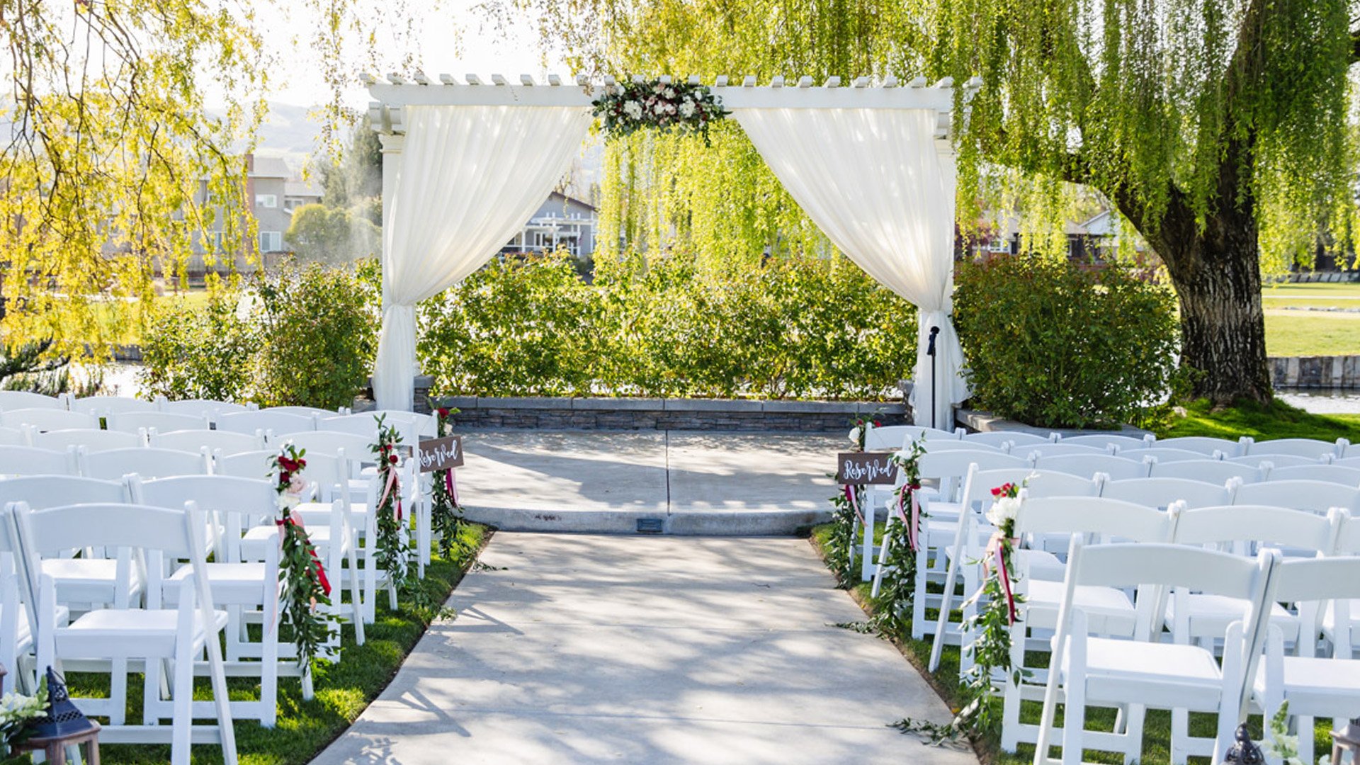 Outdoor Wedding Ceremony Setup with White Pergola Arch, Floral Decorations, and Garden Chairs Along Paved Aisle Under Weeping Willow Trees-1