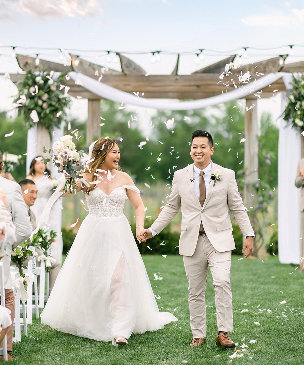 Just married couple walking down the aisle at Spinning Leaf's garden ceremony space, showered in white petals, bride in off-shoulder lace gown with blush bouquet, groom in cream suit, under wooden pergola with white draping and floral arrangements