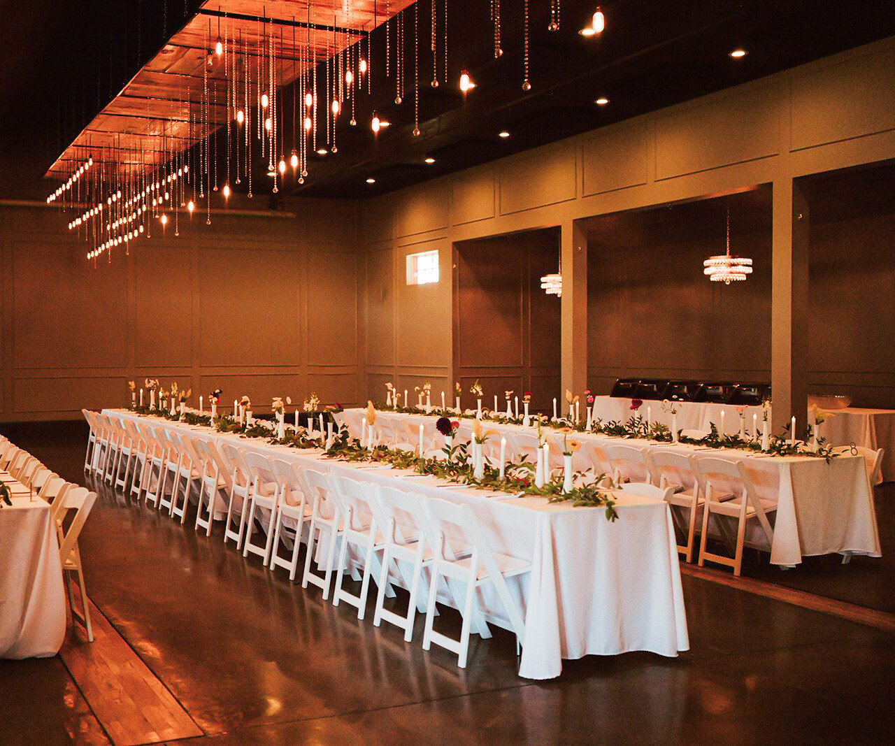 Elegant long banquet table setup in Spinning Leaf's Grand Hall, featuring cascading Edison bulb chandeliers, white tablecloths, greenery runners, and taper candles with white folding chairs