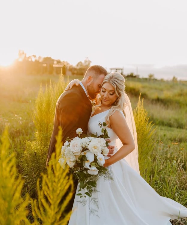 Bride and groom embracing during sunset at Spinning Leaf, surrounded by golden wild grasses, bride in white ballgown holding white rose bouquet, groom in black suit