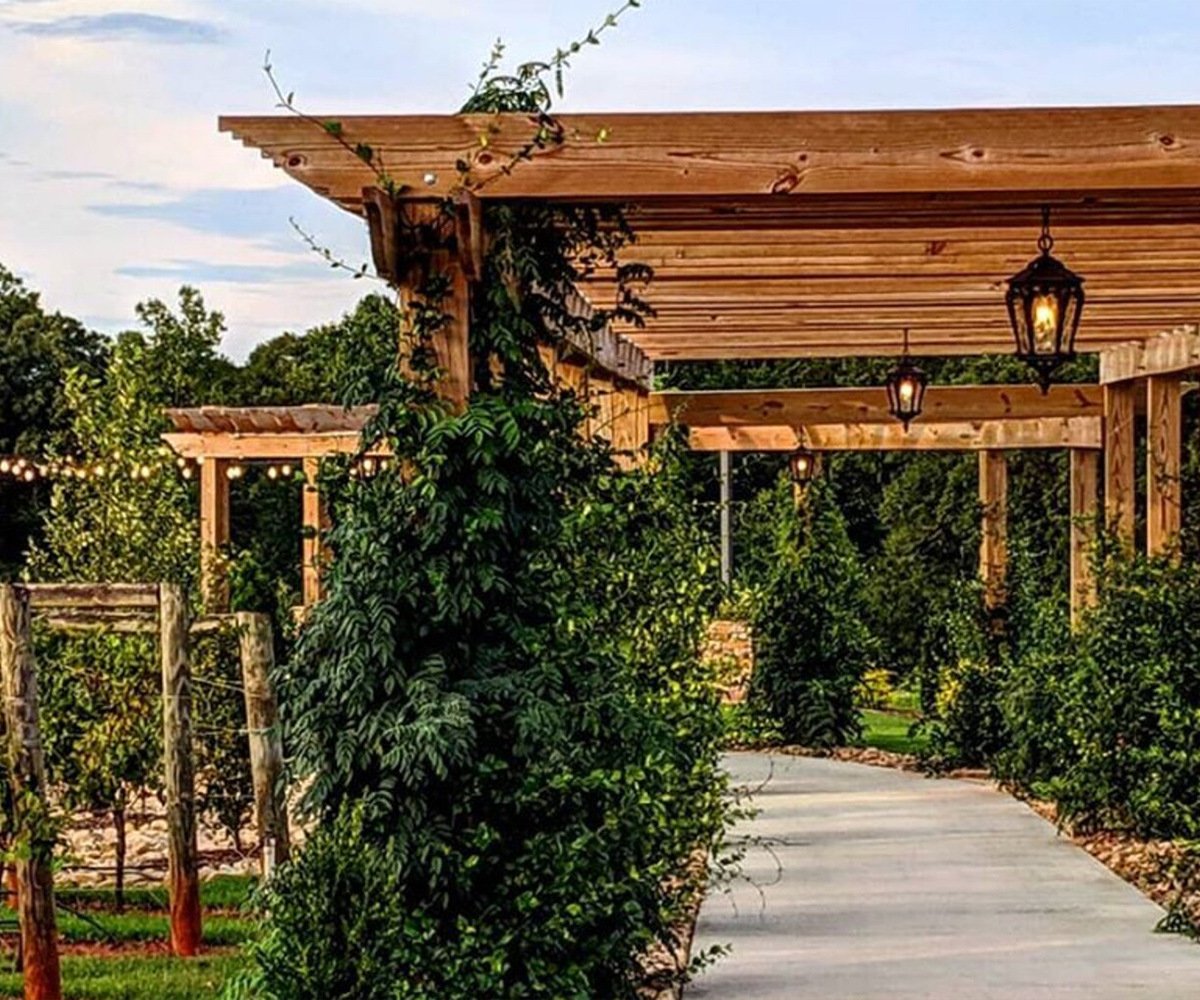 Romantic garden pathway at Spinning Leaf venue featuring wooden pergolas with climbing vines, hanging lanterns, and stone walls, creating a lush natural corridor perfect for wedding ceremonies