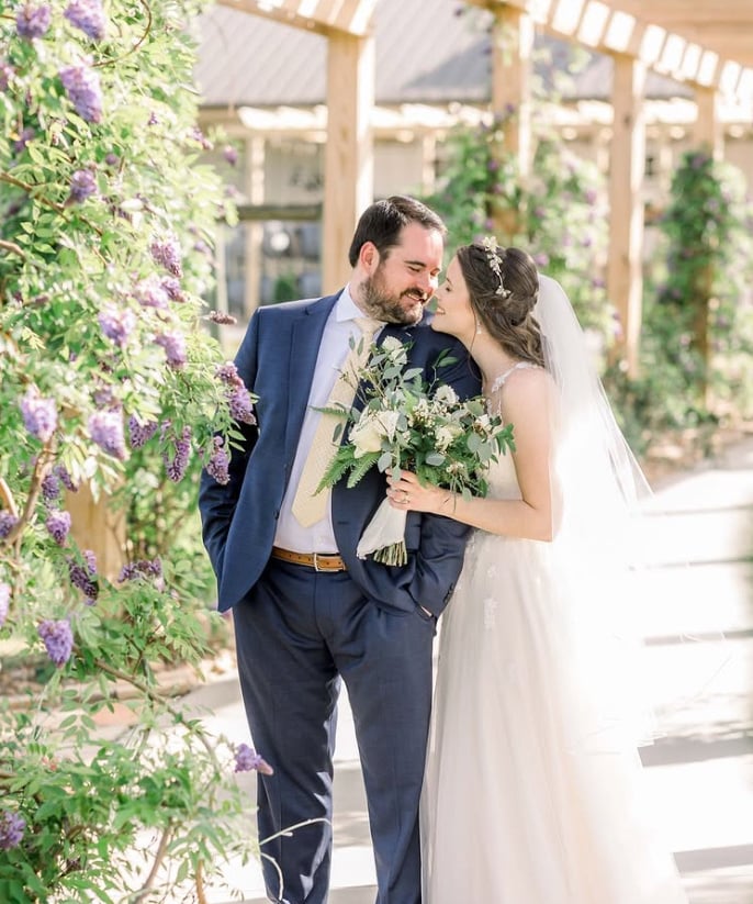 Newlyweds sharing an intimate moment under wooden pergola with blooming purple wisteria, bride in white gown holding greenery bouquet, groom in navy suit at Spinning Leaf's garden