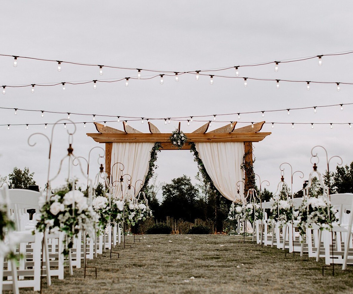 Outdoor ceremony space at Spinning Leaf featuring wooden pergola with white draping and greenery, white garden chairs lining the aisle with shepherd's hooks holding white floral arrangements, and overhead market lights creating a romantic atmosphere