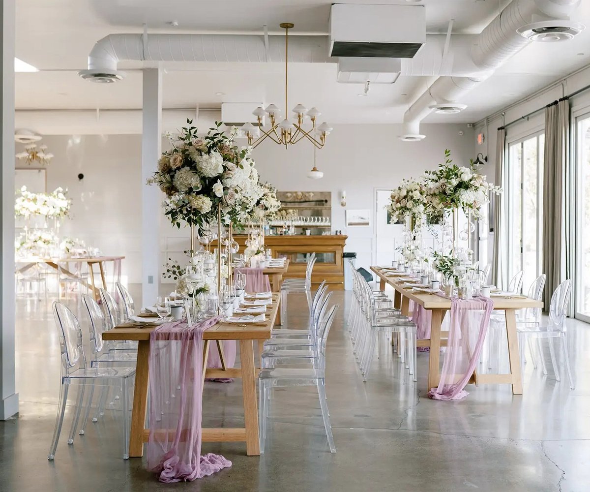 Luxury reception setup at The Harper with ghost chairs, wood farmhouse tables, blush chiffon runners and towering white floral centerpieces