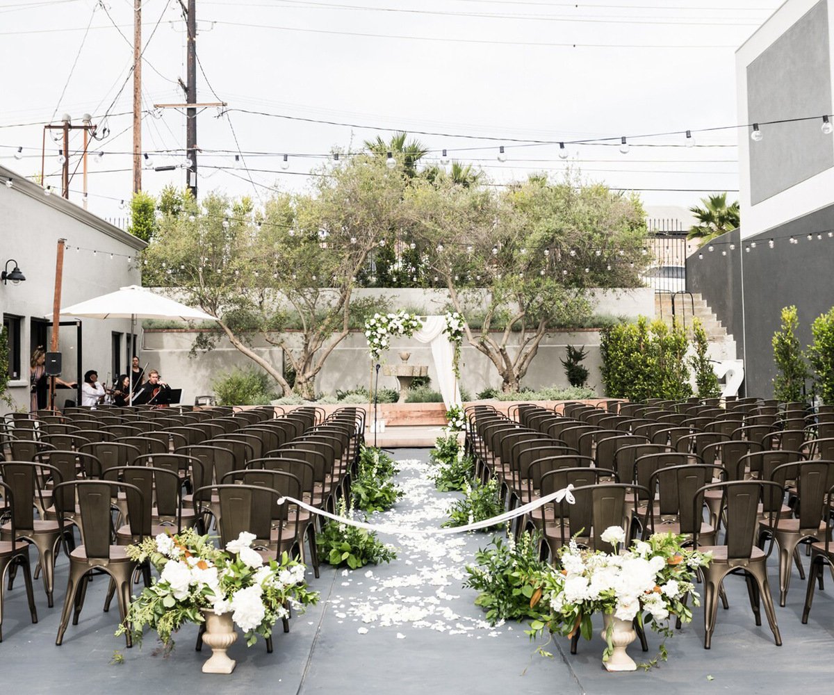 The Harper's outdoor courtyard ceremony setup with string lights, olive trees, white rose aisle markers, and metal chairs surrounded by modern architecture in Costa Mesa