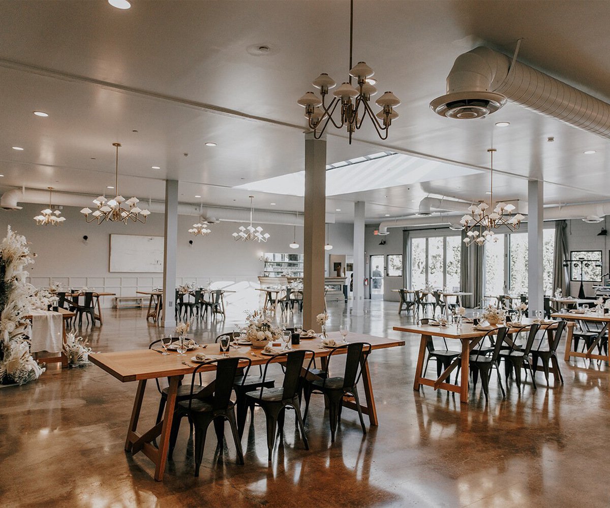 Modern industrial reception hall at The Harper with brass chandeliers, metal chairs, wood tables, and decorative dried white florals