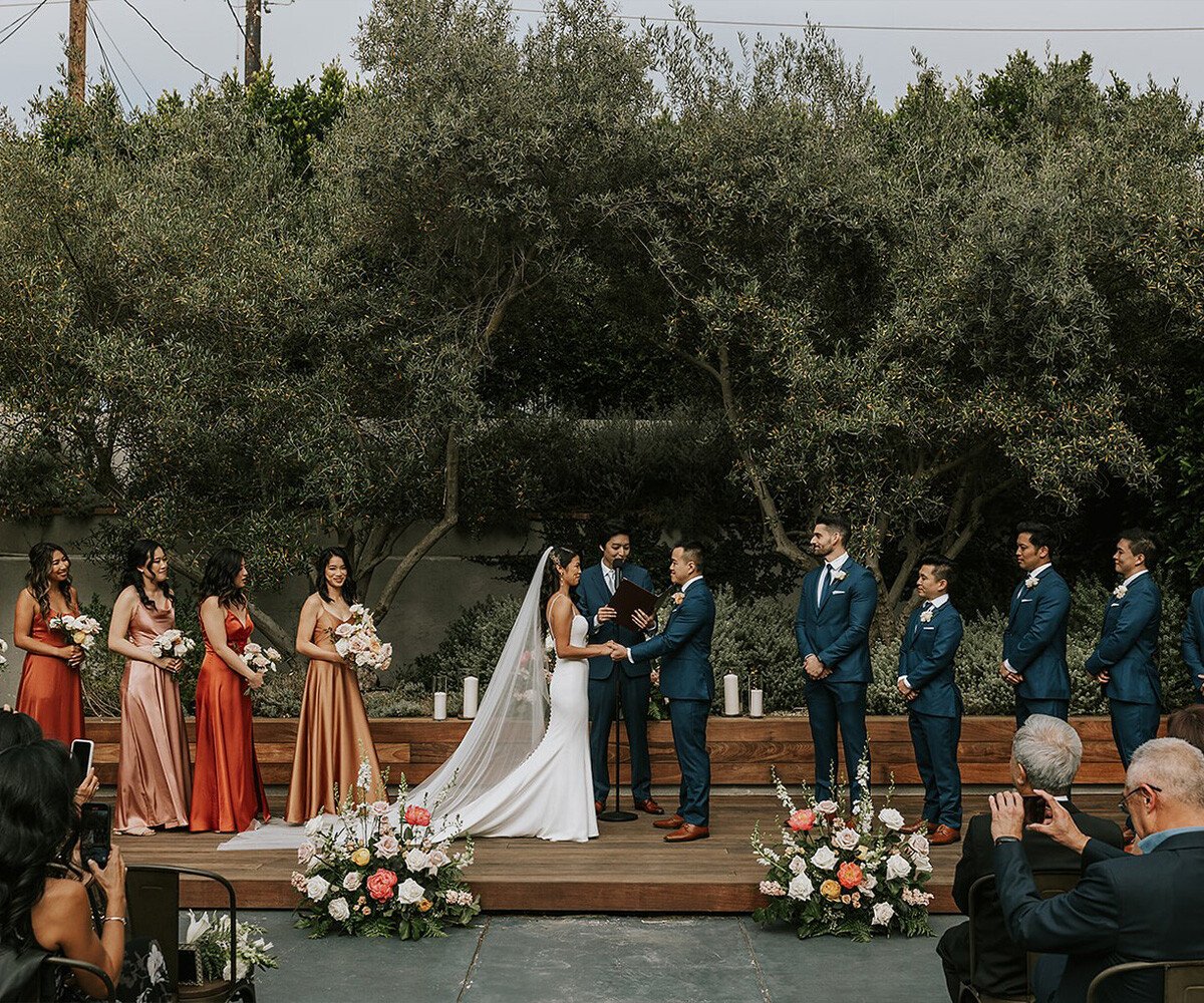 Wedding ceremony at The Harper featuring bridal party in rust and blush dresses, navy suits, olive tree backdrop and wooden stage