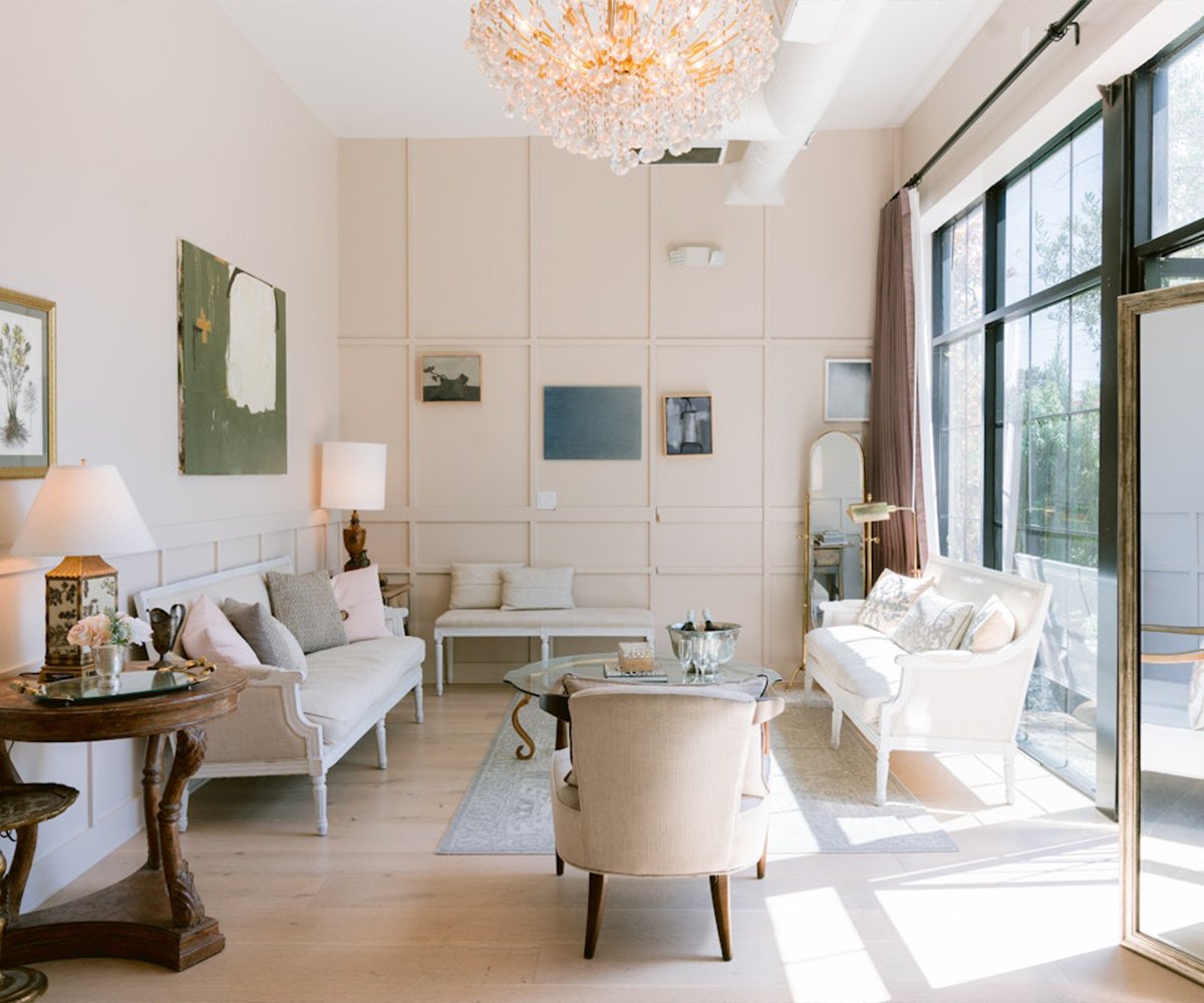 The Sunroom bridal suite at The Harper featuring crystal chandelier, all-white French provincial furniture, natural light from floor-to-ceiling windows, and modern wall paneling