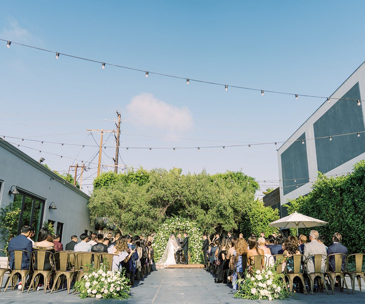 The Harper's outdoor courtyard during afternoon ceremony with market lights, floral-adorned arch, and gold metal chairs