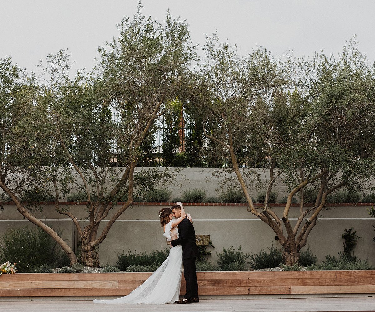 Wedding ceremony at The Harper featuring bridal party in rust and blush dresses, navy suits, olive tree backdrop and wooden stage