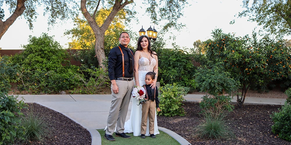 The wedding couple and their young son standing together outdoors at Lindsay Grove, surrounded by greenery and trees. The bride holds a bouquet while the groom and child, both in matching black and tan outfits, stand proudly by her side.