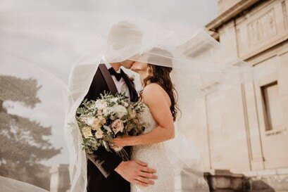 Bride and groom kissing under a wedding veil, holding a bouquet outside a historical building